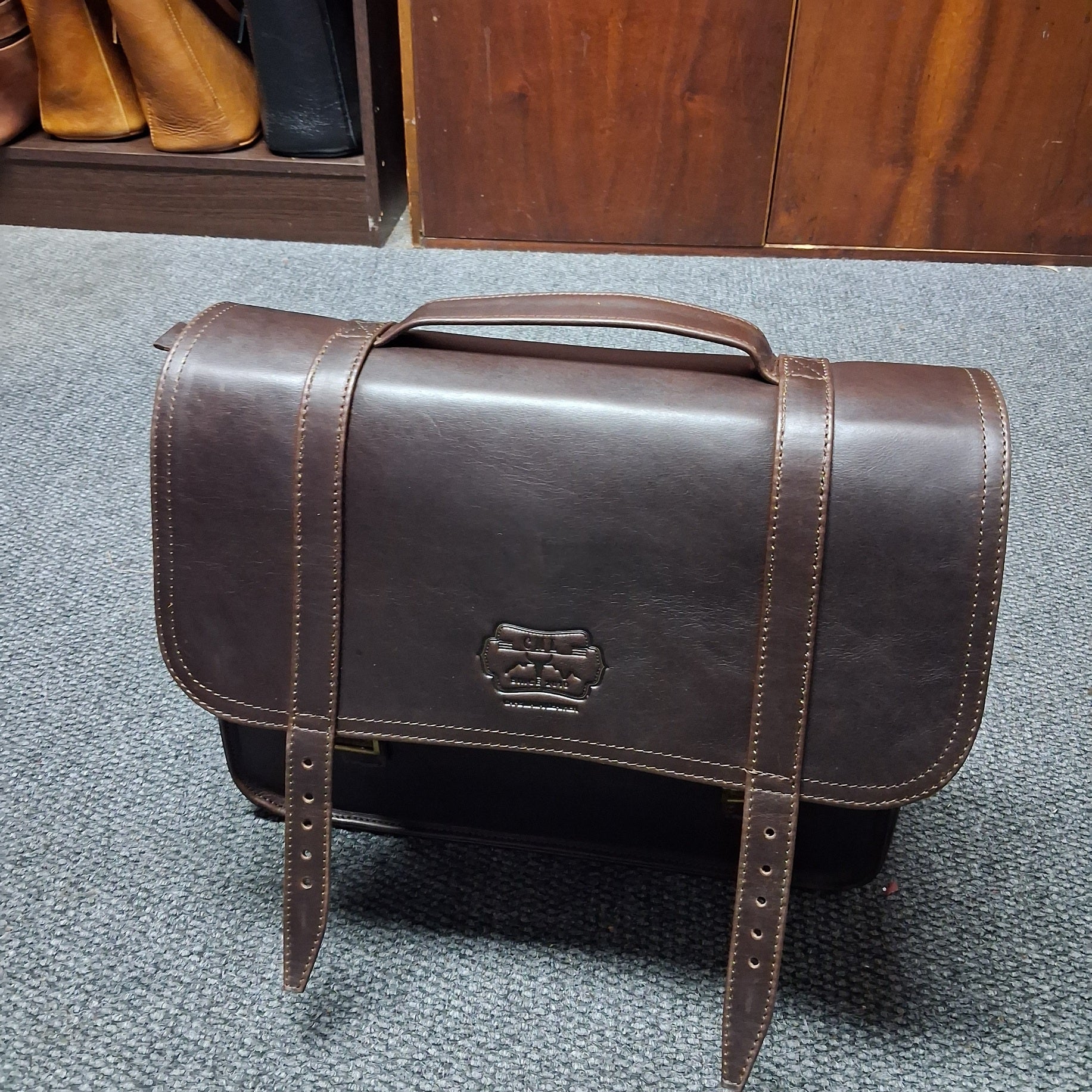 A dark brown leather briefcase or satchel with a top handle and front strap closure, photographed standing on a gray carpeted floor in front of a wooden cabinet.