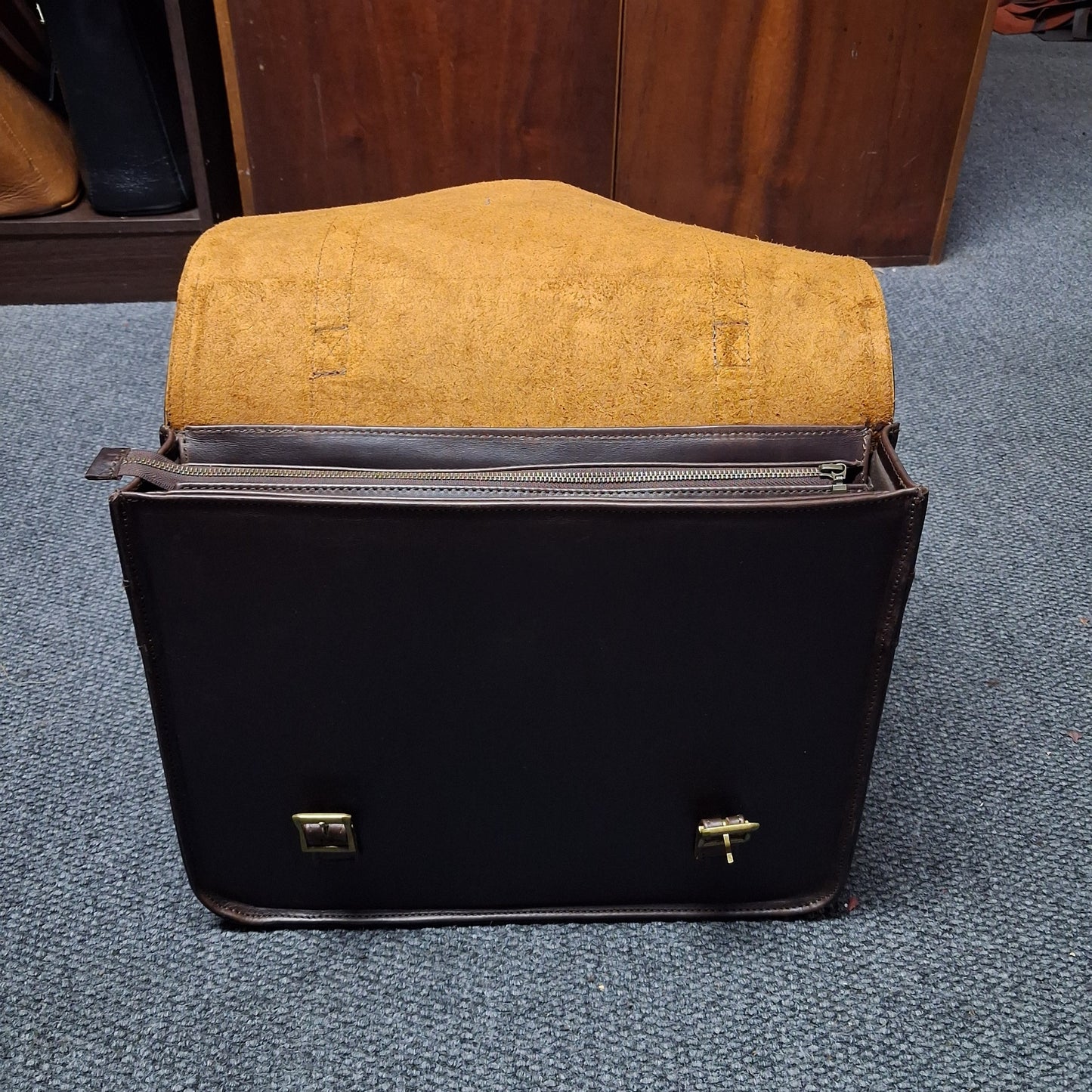 A black leather briefcase or satchel with gold-tone hardware, shown open with a tan suede or leather interior lining. The briefcase sits on a blue-gray carpeted floor in front of a wooden cabinet.