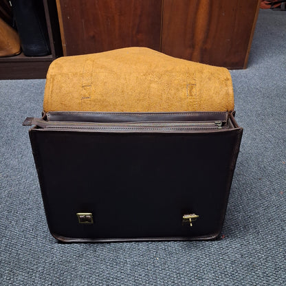 A black leather briefcase or satchel with gold-tone hardware, shown open with a tan suede or leather interior lining. The briefcase sits on a blue-gray carpeted floor in front of a wooden cabinet.