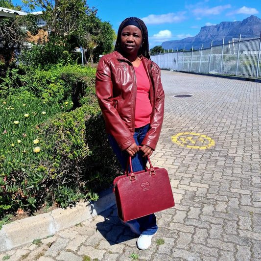 a cherry red Cape designer Laptop bag handbag with gold accents on a white background on a paved path with greenery and mountains in the background