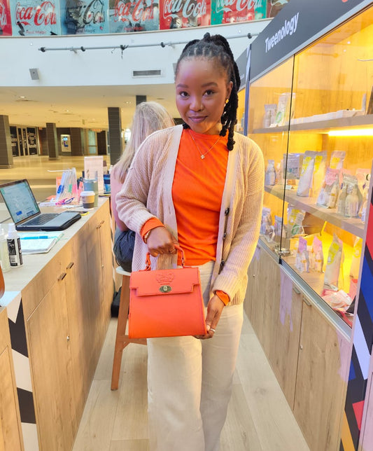 A woman holding Lawrene designer bag in orange colour in a store setting with shelves and products in the background.