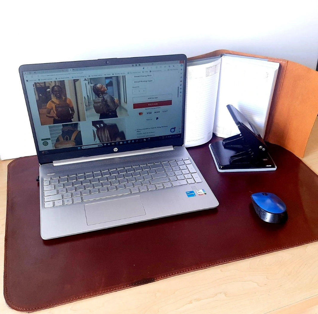 A brown leather laptop mat is displayed on a desk with a laptop on top of it, along with a blue pen and a coiled cable.