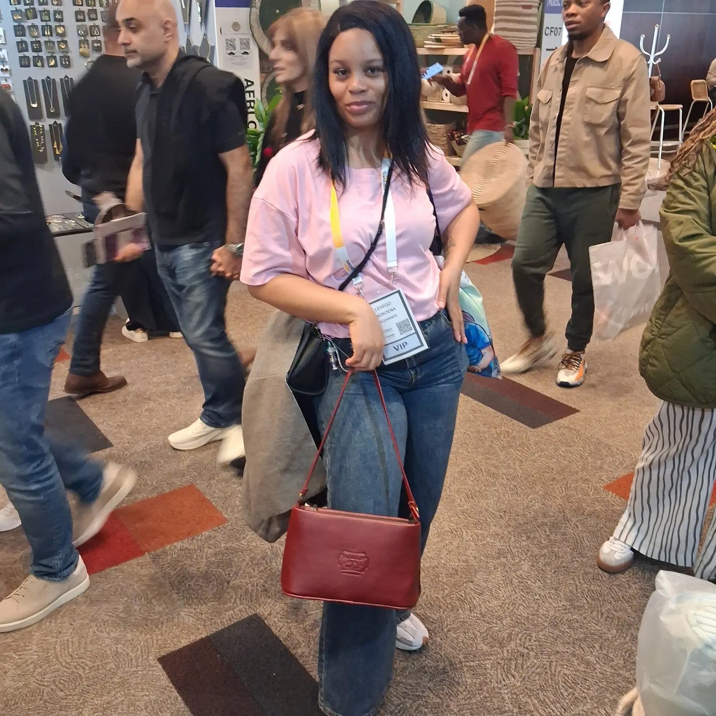 A woman at a busy indoor event or convention wearing a light pink t-shirt, blue jeans, and holding a cherry red Lucy mini shoulder bag with a gold-tone logo. She is surrounded by other people in a crowded hall with carpeted floors and booths in the background.