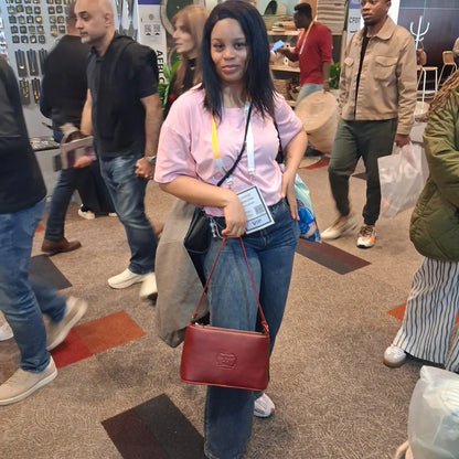 A woman at a busy indoor event or convention wearing a light pink t-shirt, blue jeans, and holding a cherry red Lucy mini shoulder bag with a gold-tone logo. She is surrounded by other people in a crowded hall with carpeted floors and booths in the background.