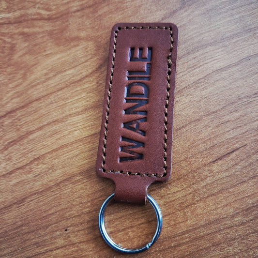 A brown leather keychain with the name 'WANDILE' embossed vertically, attached to a silver metal split ring, resting on a wooden surface.