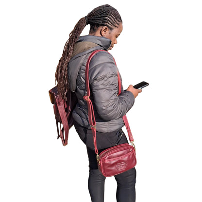 A woman with long braids stands outdoors wearing a dark gray puffer jacket, black pants, and a dark red Lady X leather crossbody bag with a matching backpack. She is looking down at a smartphone.