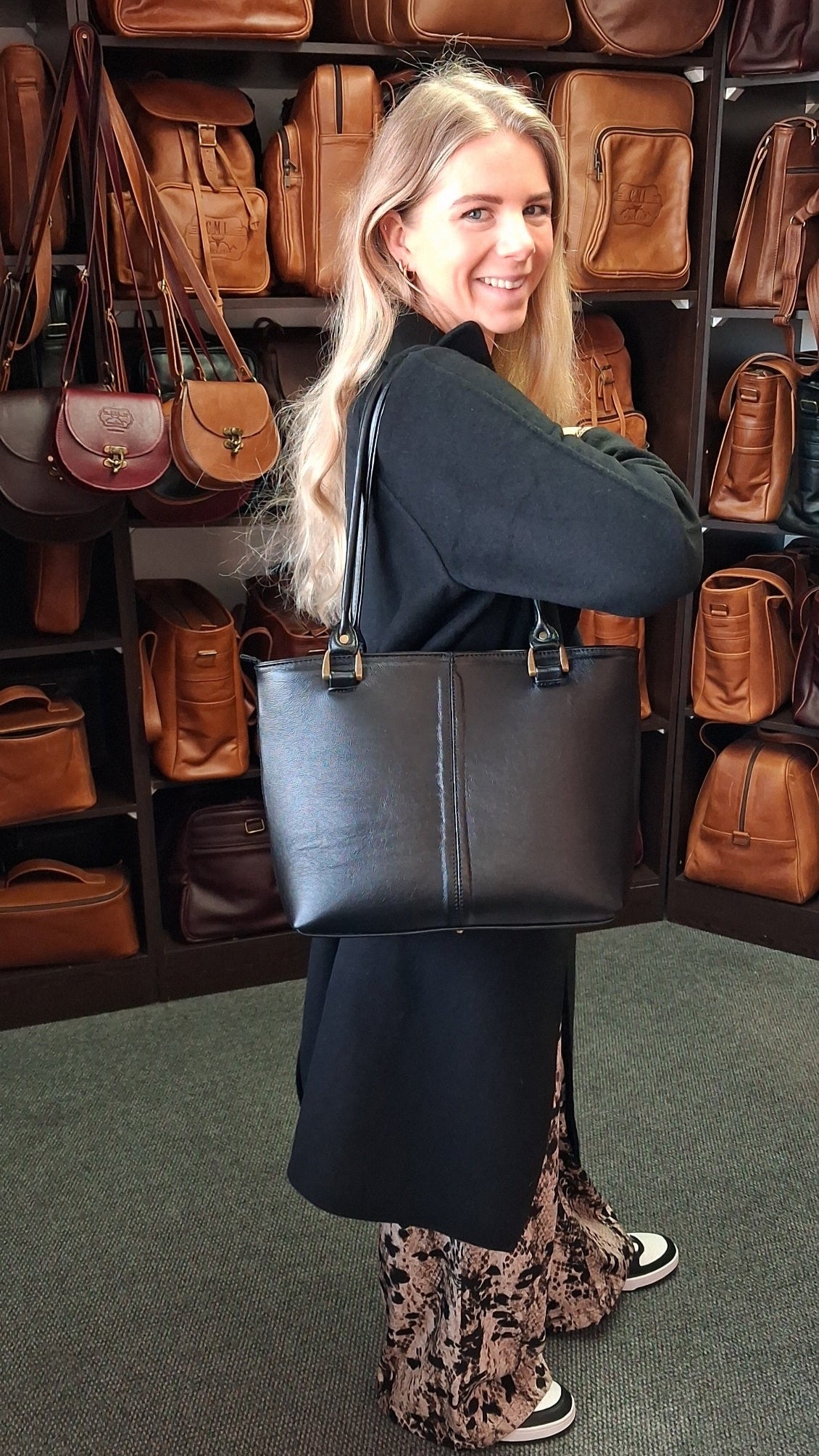 Woman holding a black Michelle leather tote bag   in front of a display of brown leather bags at Cape Masai Leather.
